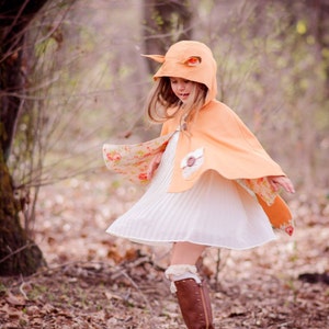 Puede incluir: Una niña pequeña con un vestido blanco y una capa con capucha de color melocotón con orejas de zorro. La capa tiene un estampado floral en el interior y un cierre de botón marrón. La niña está de pie en un área boscosa con hojas caídas en el suelo.