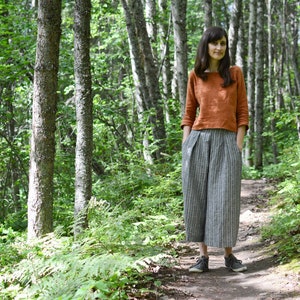 May include: A woman wearing a rust-colored linen top and striped linen pants stands on a path in a forest.