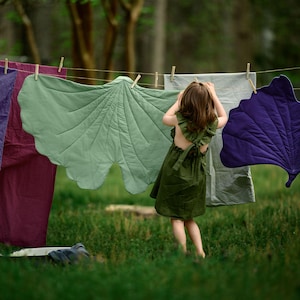 Pode incluir: Uma criança com um vestido verde está perto de um varal com tapetes acolchoados em forma de folha em tons de verde, roxo e cinza. Os tapetes são presos à corda com prendedores de roupa de madeira, criando uma cena ao ar livre lúdica.