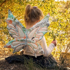 Peut inclure: Une jeune fille portant une chemise blanche et une jupe noire avec un grand costume d'ailes de papillon en tissu à motif floral. Les ailes sont bleu clair, blanc et rose avec une bordure vert clair.
