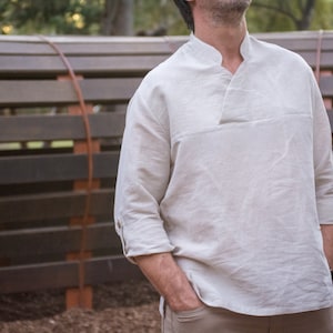 May include: A light beige linen shirt with a mandarin collar and long sleeves. The shirt has a relaxed fit and a slightly longer length.