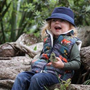 May include: A young child wearing a blue and white patterned waistcoat with a blue hat, sitting on a log in a woodland setting. The waistcoat features a whimsical woodland design with foxes, trees, and flowers.