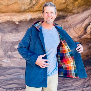 May include: A blue corduroy jacket with a plaid lining. The jacket has a hood and a button closure. The model is wearing a grey t-shirt and beige pants.