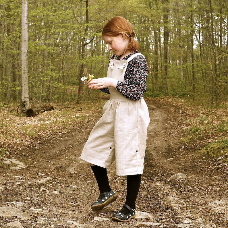 May include: A young person wearing a beige overall dress with a floral print shirt and black shoes stands on a dirt path in a forest. They are holding a small bouquet of flowers.