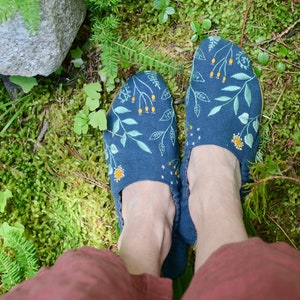 May include: Dark blue slippers with embroidered floral and leaf designs in light green, yellow, and white. The slippers are set on a bed of green moss and foliage.