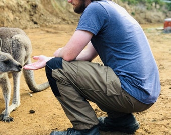 May include: A man wearing a blue t-shirt and brown pants is crouching down and looking at a grey kangaroo. The kangaroo is standing on a dirt path.