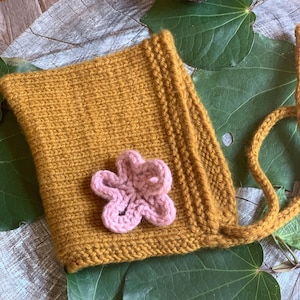 May include: A mustard yellow knitted baby hat with a pink crocheted flower embellishment and a long tie.