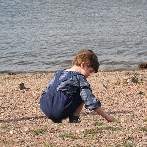 May include: A young child wearing a blue and white striped shirt and blue overalls is crouching on a pebbled beach. The child is looking down at the ground, possibly picking up a pebble.