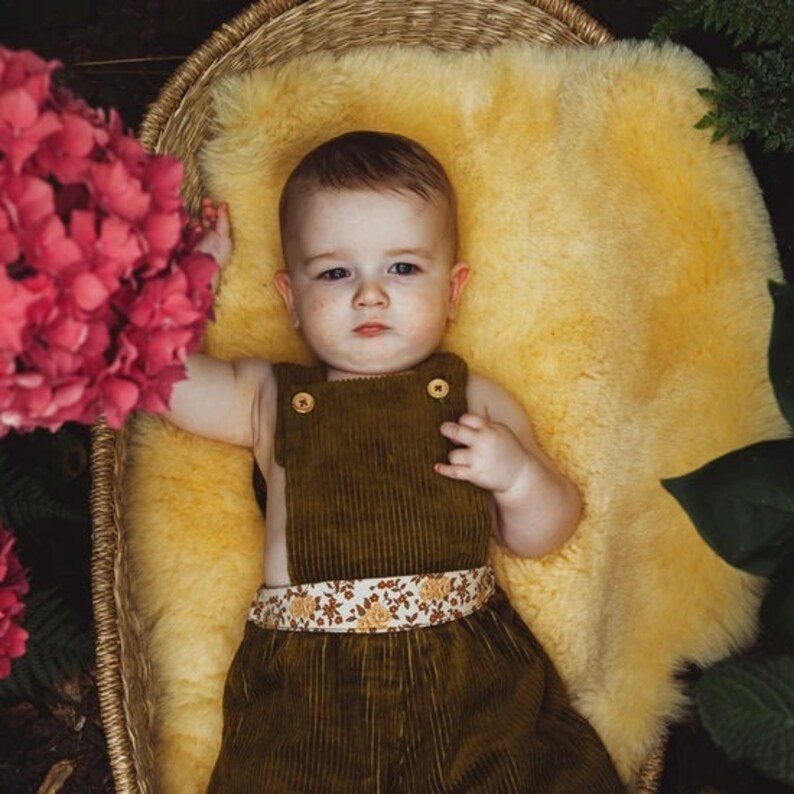 May include: A baby wearing a brown corduroy overall with a floral print waistband, lying on a yellow sheepskin rug in a wicker basket.