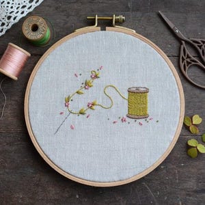May include: An embroidery hoop with a floral design and a spool of yellow thread. The embroidery features a delicate branch with pink flowers and a yellow thread spool. Additional spools of thread and scissors are visible.