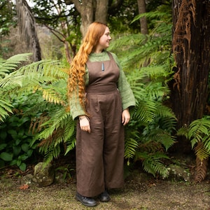 May include: A person wearing a brown jumpsuit with a green sweater underneath, standing in a lush green forest. The jumpsuit has a bib-style top and wide legs. The person has long red hair.