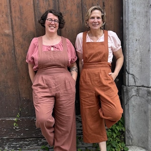 Op de afbeelding: Twee vrouwen in bijpassende jumpsuits. De ene is stoffig roze, de andere verbrand oranje. Beide hebben een vierkante halslijn en wijde pijpen. Ze staan voor een houten deur en een stenen muur. De jumpsuits zijn van linnen.
