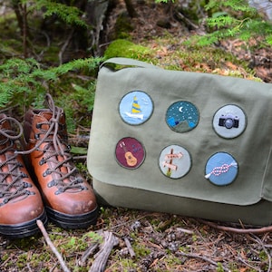 Puede incluir: Una bolsa de mensajero de lona verde con cierre de solapa y una correa larga ajustable. La bolsa tiene siete parches bordados con varios diseños, que incluyen un velero, una luna, una cámara, una guitarra, un poste indicador y un pájaro.