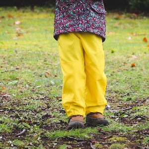May include: A person wearing yellow waterproof pants and brown boots stands on grass. The pants have reinforced knees and are paired with a floral patterned jacket. The image is taken outdoors.