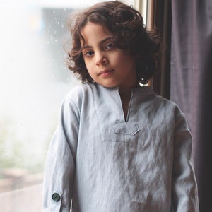 May include: A young person with curly brown hair wearing a light blue long-sleeved shirt with a V-neck and a small patch pocket on the chest.