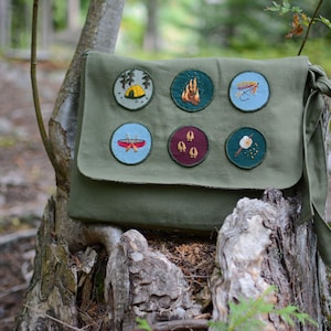 May include: A green canvas messenger bag with six embroidered patches. The patches feature a tent, a campfire, a fly fishing lure, a canoe, a footprint, and a marshmallow on a stick.