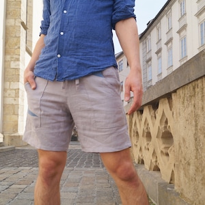 Puede incluir: Un hombre con una camisa de lino azul claro y pantalones cortos de lino beige con cintura de cordón. Los pantalones cortos tienen un diseño azul en el lateral.