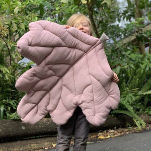 May include: A pink quilted leaf-shaped play mat with a child holding it up. The mat is made of soft fabric and has a quilted design.