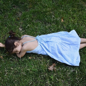 May include: A young person wearing a light blue sleeveless dress lies on their stomach on a grassy lawn. The dress has a tie at the back of the neck.