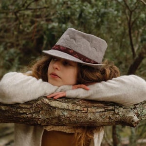 May include: A young person wearing a gray felt fedora with a floral ribbon, resting their chin on a tree branch. They are wearing a light gray sweater and have long brown hair.