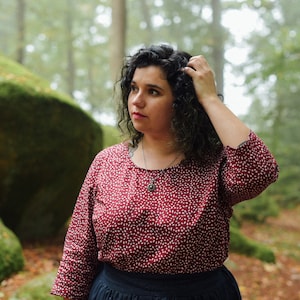 Könnte beinhalten: Eine Frau mit lockigem, dunklem Haar trägt eine rot-weiß gemusterte Bluse mit einer silbernen Halskette. Sie steht in einem Wald mit moosbewachsenen Felsen im Hintergrund.