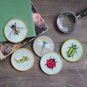 May include: Embroidered insect patches including a bee, dragonfly, grasshopper, ladybug, and beetle. Each round patch has a detailed insect design and a contrasting border. The patches are on a wooden surface with a magnifying glass and books.