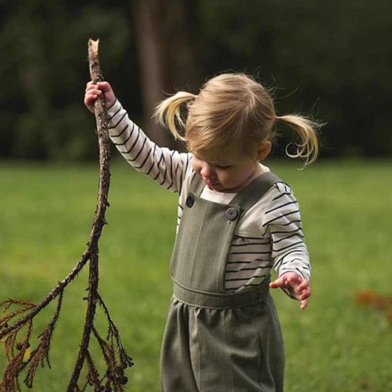 May include: A young child wearing a green overall dress with a white and black striped long-sleeved shirt. The child has pigtails and is holding a branch in their hand.