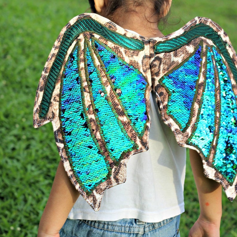 Peut inclure: Une paire d'ailes en sequins turquoise et vertes avec un dos en tissu imprim&eacute; l&eacute;opard brun. Les ailes sont attach&eacute;es &agrave; une chemise blanche.
