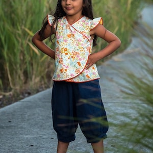 May include: A young person wearing a white and orange floral print wrap top with ruffled sleeves and navy blue pants.