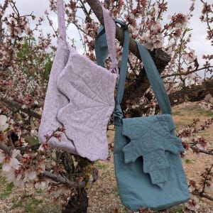 Puede incluir: Dos bolsos de hombro de tela con aplicaciones en forma de hoja cuelgan de una rama de árbol. Un bolso es de color lila claro con un estampado de lunares blancos, y el otro es de color verde azulado. Accesorios hechos a mano.