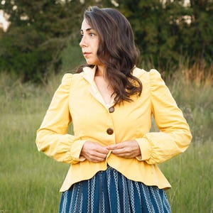 Puede incluir: Chaqueta amarilla con cuello claro y botones marrones. La chaqueta tiene mangas abullonadas y se combina con una falda a rayas azules y blancas. La persona está de pie en un campo de hierba.