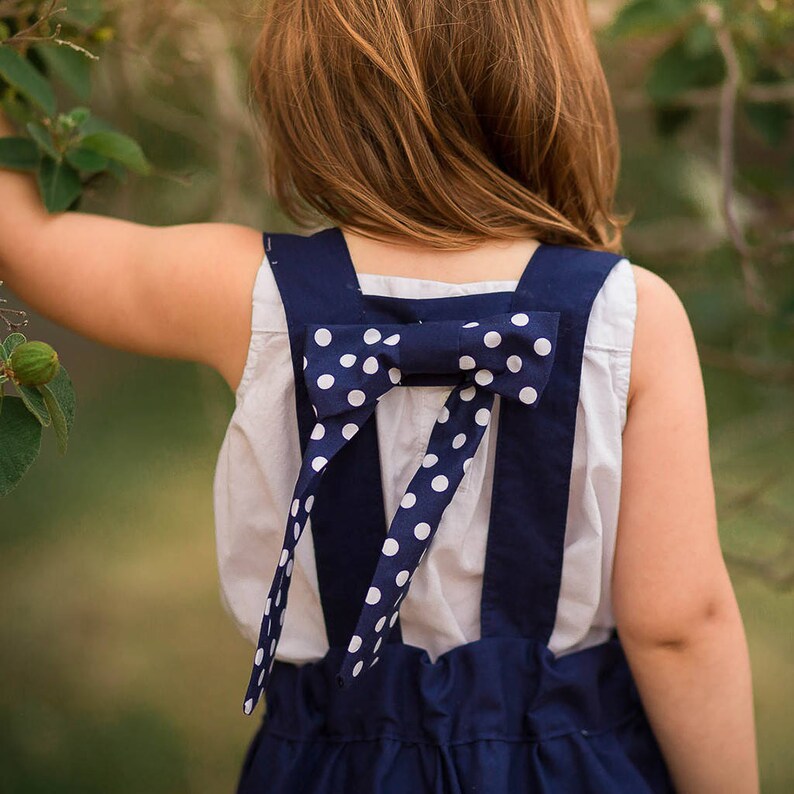 May include: A white sleeveless top with a navy blue overall dress. The straps of the dress are tied in a bow at the back with white polka dots.