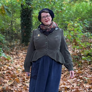 Puede incluir: Una chaqueta verde oliva con cintura peplum y una falda azul marino. La persona lleva una boina negra, gafas y una bufanda estampada. Un pequeño broche dorado está en la chaqueta.