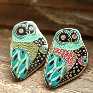 May include: Two enamel pin badges featuring colorful owl designs. The owls are depicted with intricate patterns in shades of turquoise, green, red, and black. The pins are perfect for adding a touch of whimsy to bags, jackets, or hats.