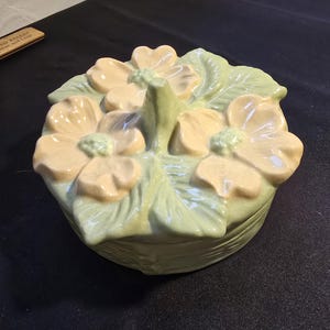 May include: A ceramic floral-shaped trinket box with a lid. The box is light green with yellow and tan flower accents and green leaves. The lid has a central handle. The box is on a black surface.