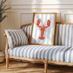 May include: A framed watercolor print of a red lobster against a blue and white striped background. The artwork is displayed on a striped blue and white sofa with a matching pillow. The frame is a light brown color.