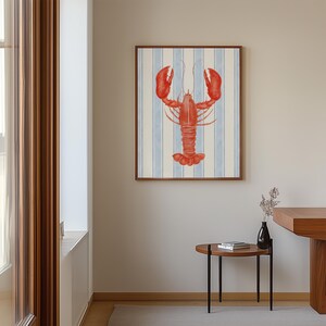 May include: A framed art print featuring a red lobster illustration on a blue and white striped background. The artwork has a wooden frame and is displayed on a neutral-colored wall. The print adds a nautical theme to the room.