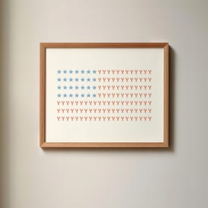 Puede incluir: Impresión artística enmarcada que presenta un diseño estilizado de la bandera estadounidense. La obra de arte utiliza estrellas azules y símbolos rojos en forma de langosta sobre un fondo crema. La impresión está en un marco de madera natural, para decoración de pared.