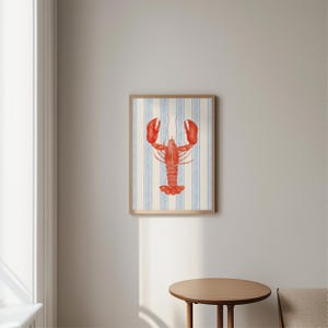 May include: A framed art print featuring a red lobster illustration. The lobster is centered on a background of vertical blue and white stripes. The frame is a light brown color, and the overall style is nautical.