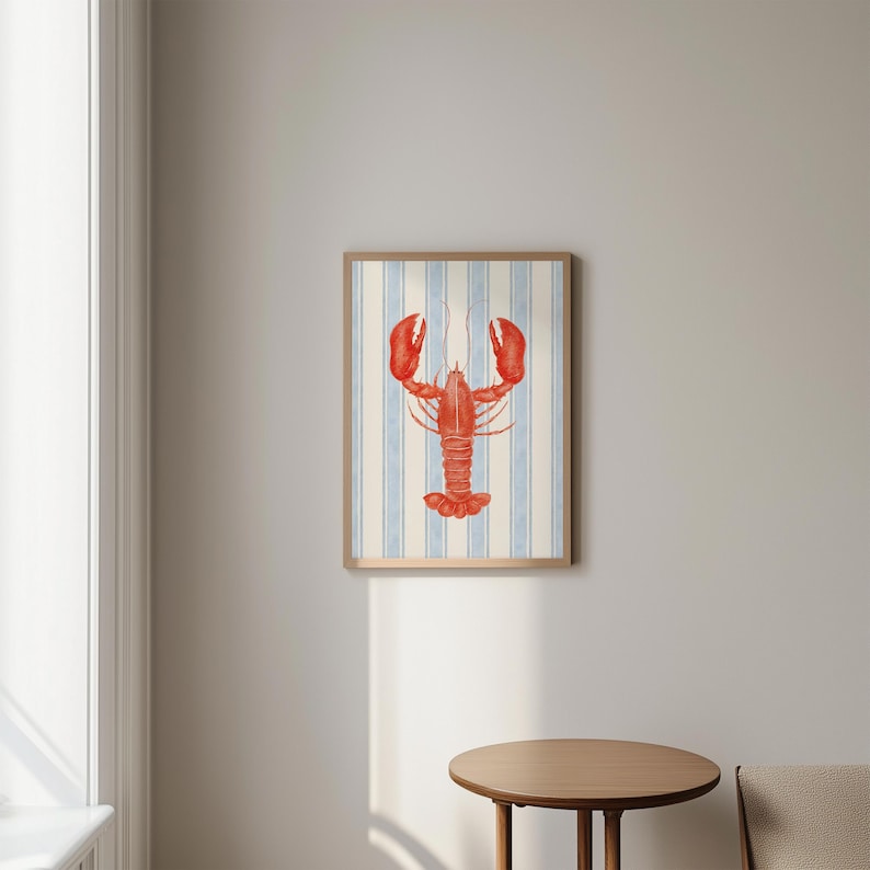 May include: A framed art print featuring a red lobster illustration. The lobster is centered on a background of vertical blue and white stripes. The frame is a light brown color, and the overall style is nautical.