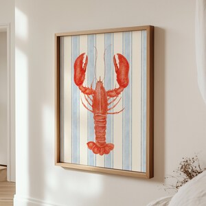 May include: A framed art print featuring a vibrant red lobster illustration. The lobster is centered against a backdrop of vertical blue and cream stripes. The artwork is encased in a natural wood frame, adding a touch of coastal charm.
