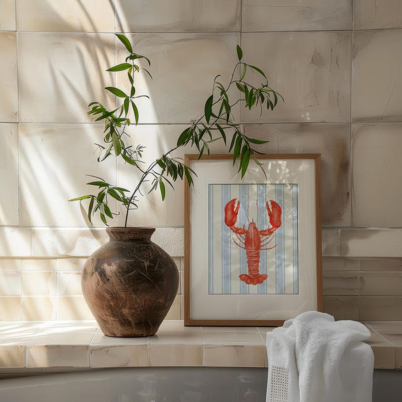 May include: A decorative still life featuring a framed lobster print with blue and white stripes, a brown vase with greenery, and a white towel. The lobster art is in a wooden frame. The vase is brown with a textured surface.