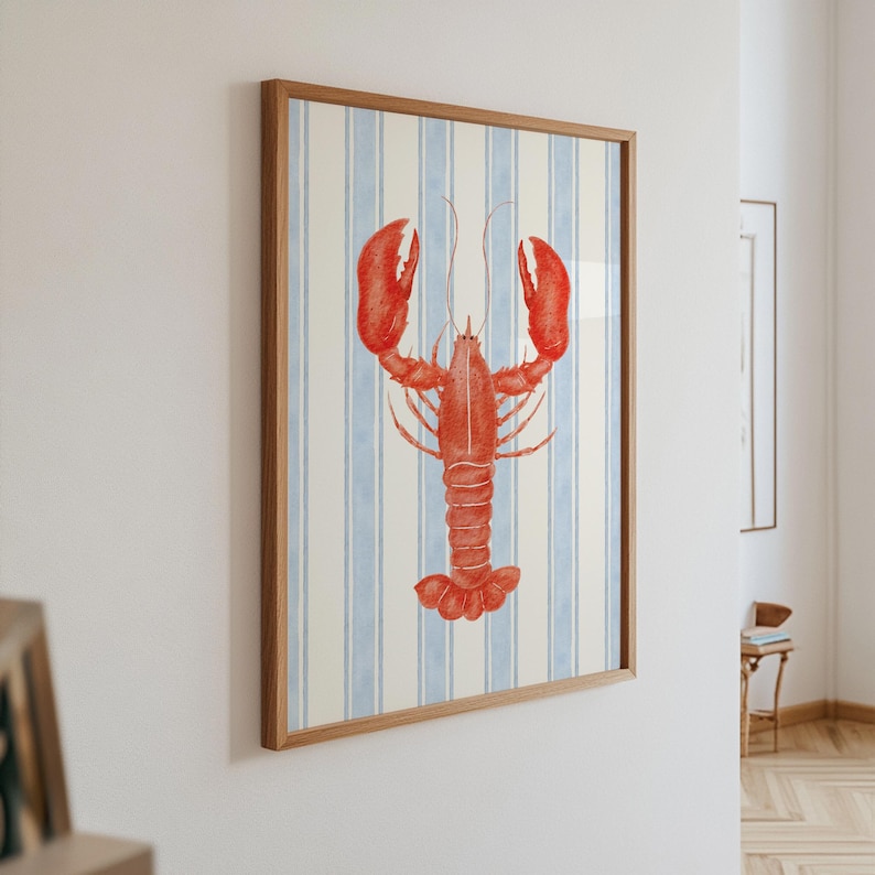 May include: A framed art print featuring a watercolor illustration of a red lobster. The lobster is centered against a background of vertical blue and white stripes. The frame is made of light-colored wood, adding a nautical theme.