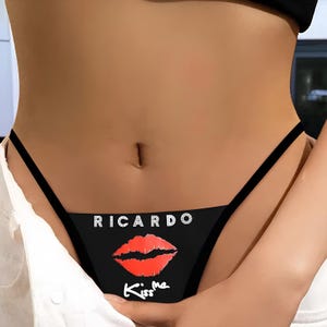 Può includere: Perizoma nero con il nome "RICARDO" in lettere argentate sopra un disegno di labbra rosse. Le parole "Kiss me" sono scritte sotto le labbra in corsivo bianco.
