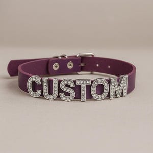 Może przedstawiać: Fioletowa skórzana obroża z napisem "CUSTOM" w srebrnych literach wysadzanych przezroczystymi kryształkami. Obroża posiada srebrną klamrę i dwa srebrne nity.
