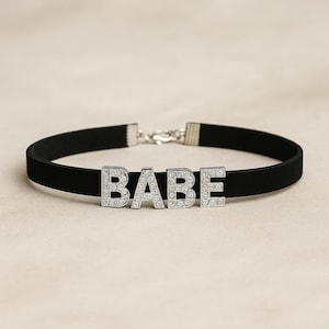 Może przedstawiać: Czarny naszyjnik typu choker z napisem "BABE" w srebrnych literach wysadzanych przezroczystymi kryształkami. Naszyjnik ma srebrne zapięcie i jest prezentowany na neutralnym tle.