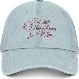 May include: A light blue denim baseball cap featuring the embroidered dark red text "I Don't Need a Reason to Wine." The cap has a six-panel construction with a curved brim, a button on top, and stitched eyelets for ventilation on each panel.