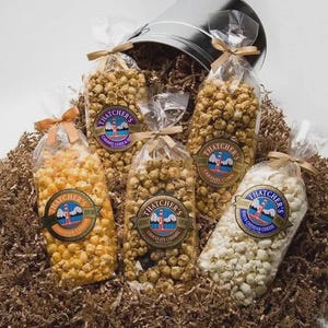 May include: An assortment of popcorn bags, including caramel corn and cheddar cheese flavors, are arranged on a bed of brown paper. Each bag is sealed with a ribbon and features a label with a lighthouse logo. A metal bucket is visible in the background.