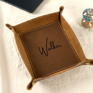 May include: A brown leather valet tray with the name "Walker" embossed in the center. The tray has a square shape with raised edges and is stitched with white thread. The corners are secured with gold-colored rivets. A laptop and decorative glass sphere are in the background.