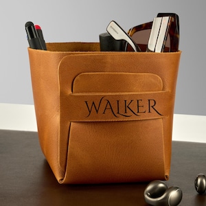 Pode incluir: Um organizador de mesa de couro castanho com o nome "WALKER" impresso na frente. O organizador contém canetas, óculos de sol e outros itens. Um caderno, uma garrafa de água e abotoaduras também são visíveis na mesa.